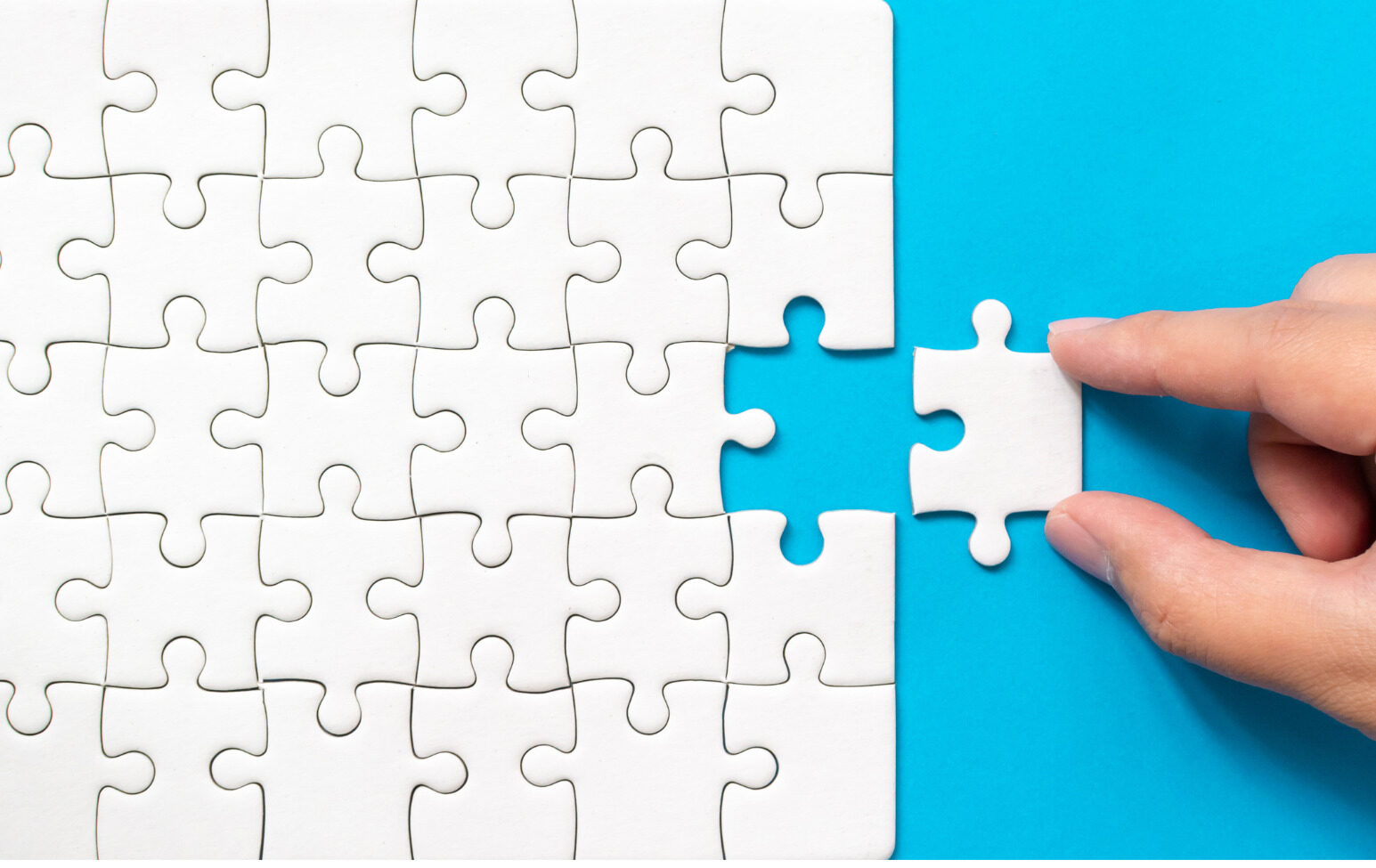 A hand placing a white puzzle piece onto a white puzzle with a bright blue background.