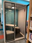 meeting booth for two people with two seats and a table inside and clear doors and opaque sides, locating inside the library